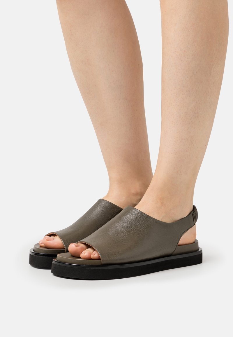 Fötter i öppna sandaler med bakrem i mörk olivgrön läder och tjocka svarta sulor på vit bakgrund.