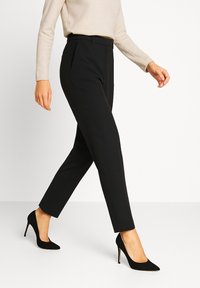 Pantaloni neri sartoriali con una vestibilità slim, caratterizzati da una texture liscia, vita media e sottili pieghe frontali, abbinati a tacchi neri.
