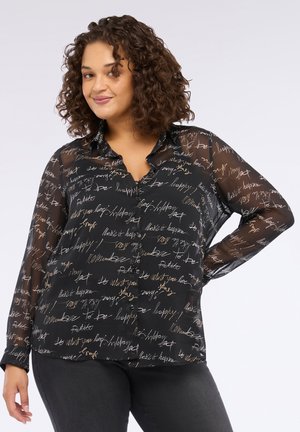 Curly-haired woman wearing a sheer black button-up blouse with white and tan handwritten text patterns, paired with dark pants.