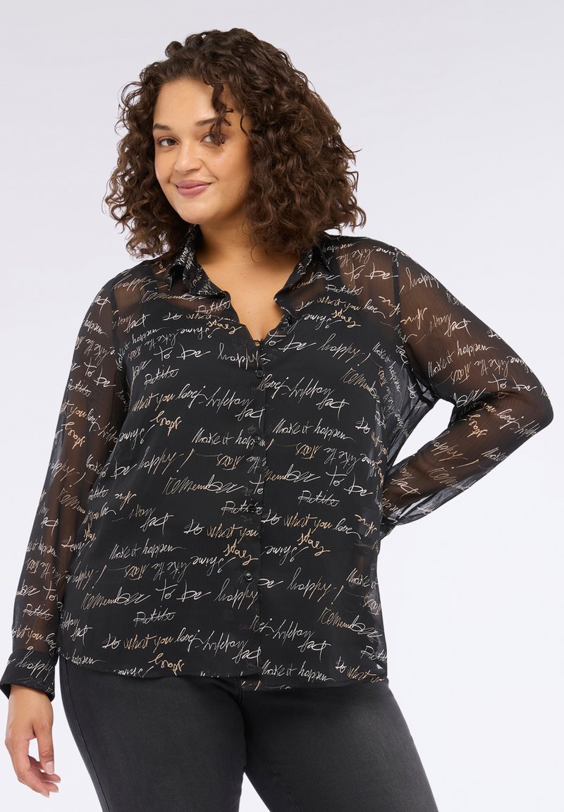Donna dai capelli ricci che indossa una blusa nera trasparente con bottoni, decorata con scritte a mano bianche e beige, abbinata a pantaloni scuri.