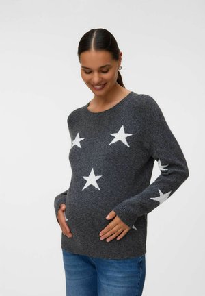 Pregnant woman in a dark gray sweater with white stars, holding her belly and smiling with head slightly bowed.