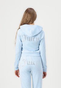Lichtblauwe velours trainingspak met een aansluitende sweatshirt met capuchon. "Juicy" in strasssteentjes op de achterkant, bijpassende broek met "Juicy" op de achterkant.