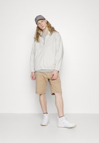 Lys grå half-zip jakke med elastisk kant, beige shorts og hvide sneakers. Grå kasket fuldender outfittet. Minimalistisk design med afslappet pasform.