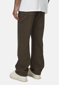 Luźne brązowe bawełniane spodnie z kieszeniami bocznymi i kontrastowym przeszyciem; noszone z białymi butami i luźną białą koszulą.