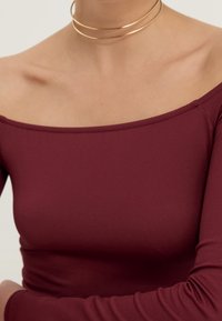 Burgundy topp utan axlar i stretchmaterial, med en figursydd silhuett och långärmar med en smidig textur. Guldhalsband som accent.