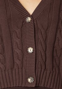 Cardigan marrón de chunky con trenzas y tres botones de carey, con escote en V, llevado por una persona.