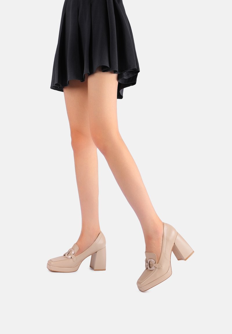 Scarpe con tacco alto beige con punta rotonda, caratterizzate da una fibbia decorativa e un tacco grosso, abbinate a una gonna nera a campana.