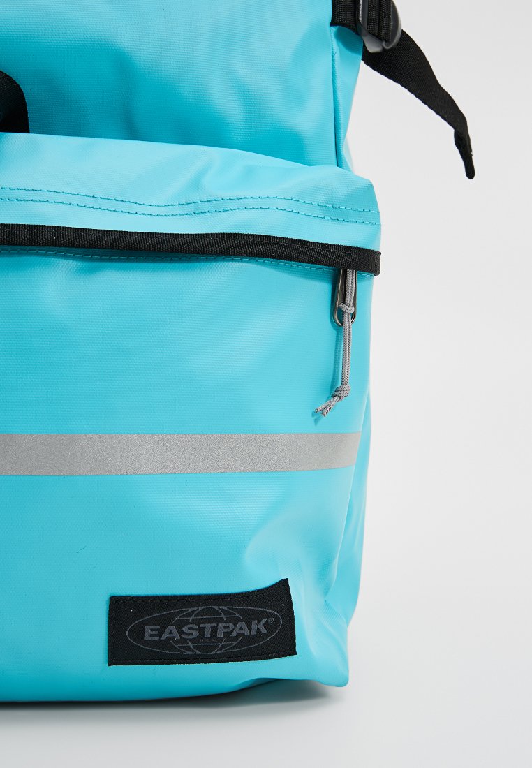 Türkise Rucksack aus robustem Material, mit einem silbernen reflektierenden Streifen, schwarzem Reißverschluss und Eastpak-Logopatch an der Unterseite.