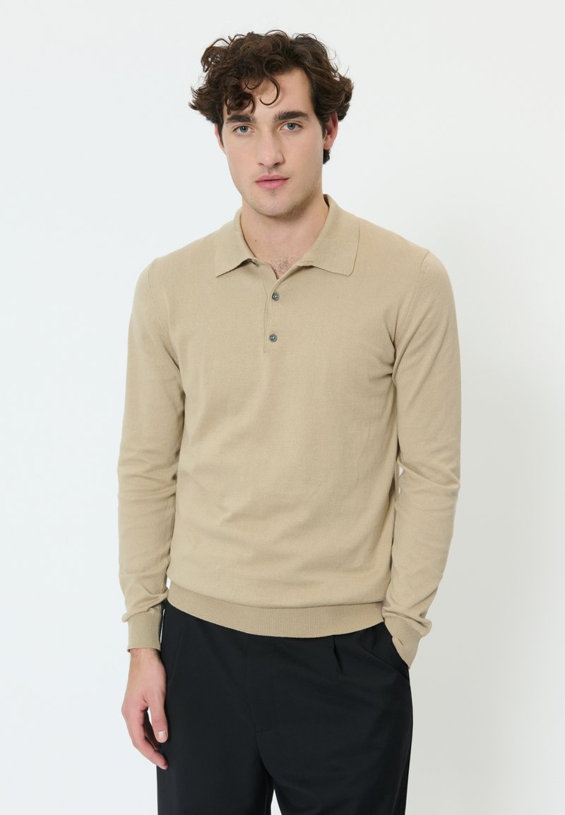 Man met krullend bruin haar, gekleed in een beige poloshirt met lange mouwen en zwarte broek, staand tegen een witte achtergrond.