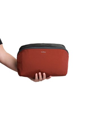 Main tenant une pochette zippée Bellroy de taille moyenne, rouge et noire, sur un fond blanc.