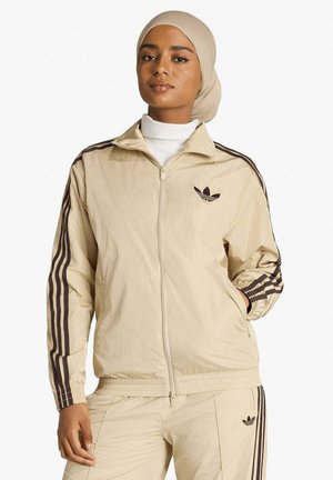 Donna che indossa una tuta Adidas beige con strisce e logo marroni, abbinata a un hijab beige, in piedi davanti a uno sfondo bianco.
