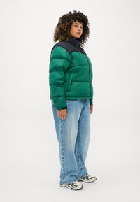 Grøn pufferjakke med sorte detaljer, lynlås foran og sidelommer. Modellen bærer lys blå vide jeans og mønstrede sneakers.