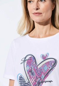 Witte t-shirt met een groot hartontwerp in kleurrijke geometrische patronen in roze, blauw en paars, verfraaid met de tekst "Spirit of Summer".
