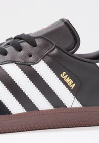 Sneaker Adidas Samba nera con strisce bianche, testo dorato "SAMBA", lacci neri e suola texture marrone, mostrato in dettaglio laterale.