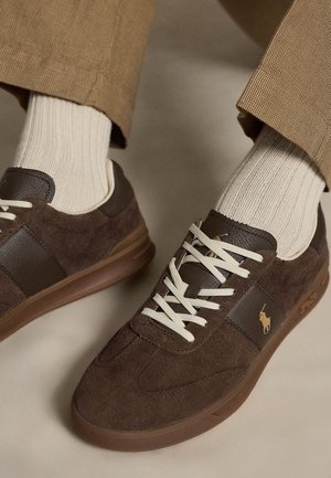 Bruine suède en leren sneakers met witte veters en een gouden logo, gedragen met beige geribbelde sokken en beige textuurbroeken.