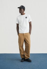 Λευκό βαμβακερό t-shirt με μαύρο λογότυπο, σε συνδυασμό με καφέ παντελόνι από καμβά και μαύρα αθλητικά παπούτσια. Το μοντέλο φοράει μαύρο καπέλο.