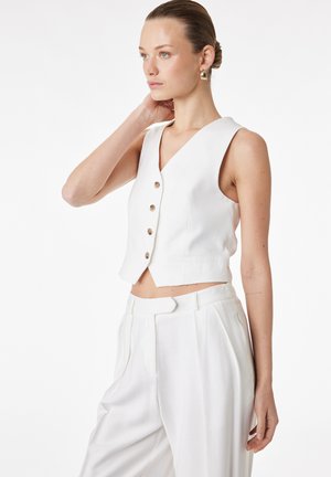 Kvinde med hår sat op i en knold, iført guldsmykker, en ærmeløs hvid vest med knapper og hvide bukser med høj talje, der kigger til siden.