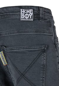 Schwarze Denim-Hose mit einem rechteckigen Logo-Tag und einem farbenfrohen Seitenetikett. Der Stoff hat eine glatte Textur mit ausgeprägten Nähdetails.