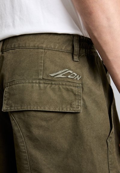 Olivgrüne Hose mit elastischem Bund, ausgestattet mit einer Gesäßtasche und einem gestickten Logo in Hellgrau auf der rechten Seite.