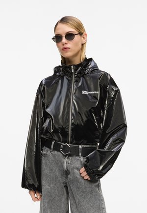 Frau trägt schwarze glänzende Kapuzenjacke mit "Karl Lagerfeld Jeans"-Logo, graue High-Waist-Jeans, schwarzen Gürtel und kleine schwarze Sonnenbrille.