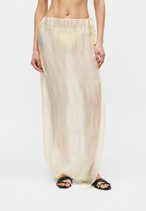 Žena v priehľadnej, bledozltej sukni dlhá po členky so postrannými viazačkami a čiernych slide sandáloch so zlatými akcentmi.