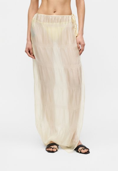 Femme portant une jupe transparente jaune pâle longueur cheville avec des liens sur les côtés et des sandales slides noires avec des accents dorés.