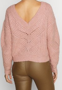 Różowy sweter na drutach z dekoltem w kształcie litery V z tyłu, zdobiony teksturowanymi wzorami i ściągaczami na mankietach. Luźny krój z opuszczonymi ramionami i skróconą długością.