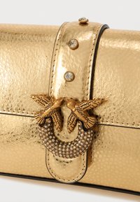 Clutch de couro dourado com textura em relevo, apresentando uma fecho decorativo com dois pássaros e realces de cristal. Acabamento preto ao longo das bordas.