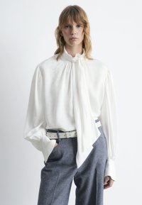 Weißes Seidenbluse mit hohem Kragen und langen Ärmeln, verziert mit einem Schleifen-Detail. Kombiniert mit grauen Hosen und einem weißen Gürtel.