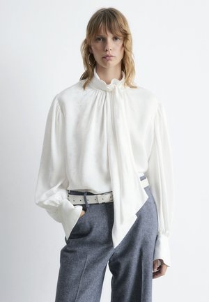 Weißes Seidenbluse mit hohem Kragen und langen Ärmeln, verziert mit einem Schleifen-Detail. Kombiniert mit grauen Hosen und einem weißen Gürtel.