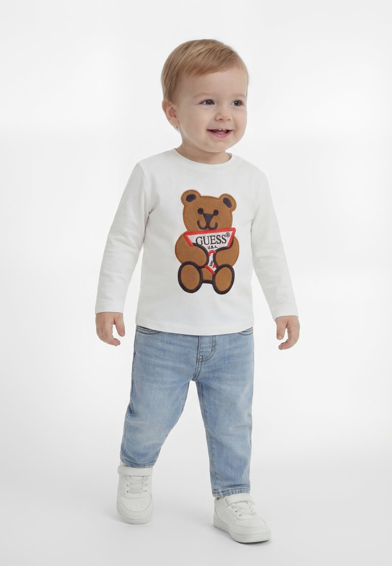 Dijete sa svijetlom kosom u bijeloj majici dugih rukava s likom medvjedića, svijetlo plavim trapericama i bijelim tenisicama, stoji i smiješi se.