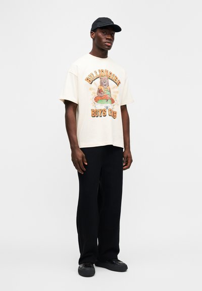 Ανοιχτό μπεζ γραφικό t-shirt με το λογότυπο "Billionaire Boys Club", συνδυασμένο με φαρδιά μαύρη παντελόνια και αθλητικά παπούτσια. Ένα μαύρο καπέλο ολοκληρώνει το ντύσιμο.