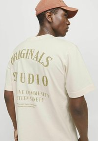 Beige katoenen t-shirt met gouden opdruk op de achterkant. De tekst bevat "ORIGINALS," "STUDIO," en aanvullende zinnen. Korte mouwen, ontspannen pasvorm.