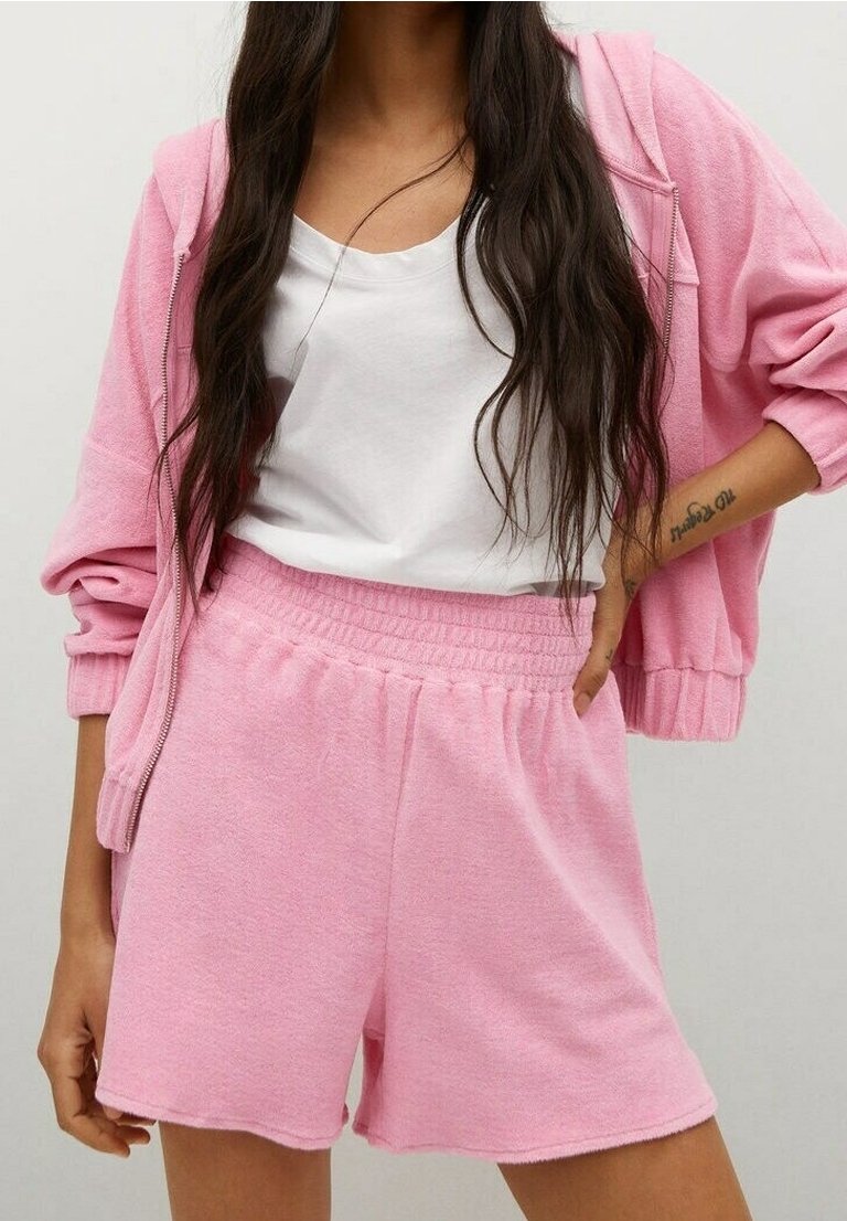 Femme portant un sweat à capuche zippé rose et un short en éponge rose assorti avec un t-shirt blanc, main posée sur la hanche.