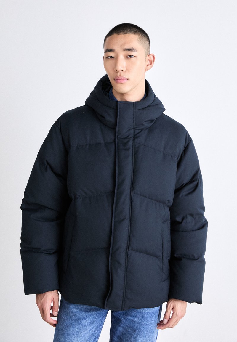 Mörk marinblå vadderad jacka med hög krage och huva, med en texturerad, quiltad design och dragkedja framtill.