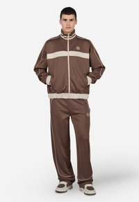 Survêtement marron en tissu lisse, avec une bande crème contrastante sur la poitrine et le long des pantalons, et des poignets élastiques.