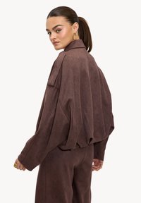 Veste surdimensionnée en daim marron avec col rabattu et épaules tombantes, offrant une coupe décontractée et une surface texturée.
