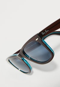 Sluneční brýle Ray-Ban s hnědým rámem a tyrkysovými akcenty, tmavými tónovanými skly a logem na straně. Elegantní geometrický design.