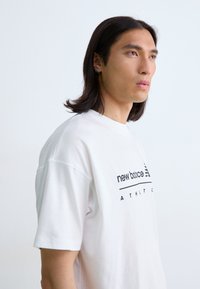 T-shirt en coton blanc avec un logo et du texte « new balance » en noir, manches courtes et coupe décontractée, porté par une personne avec de longs cheveux.