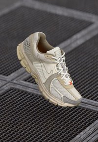 Bež i sivi Nike teniski sa bijelim vezicama i narančastim akcentom, prikazan pod kutom na metalnoj rešetkasto površini.