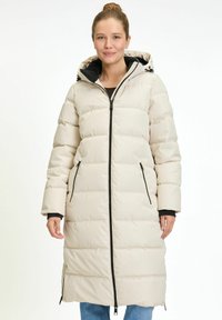 Vrouw in een lange beige pufferjack met zwarte ritsdetails, capuchon en zijventilaties met rits, gecombineerd met blauwe jeans, staand tegen een witte achtergrond.
