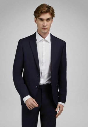 Młody mężczyzna o jasnobrązowych włosach, ubrany w ciemnogranatowy garnitur i białą koszulę, pozujący na jednolitym szarym tle.