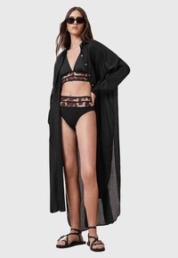 Camicia copri costume leggera in lino nera con un design lungo e aperto. Sotto, un bikini nero con fascia a motivo grafico. Sandali flat.