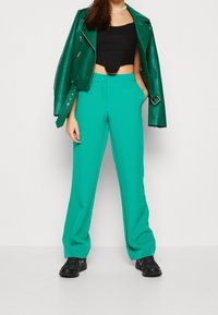 Tealfärgade vida byxor i lätt tyg, kombinerade med en svart kortärmad topp och en figurnära grön läderjacka. Svarta sneakers fulländar outfiten.