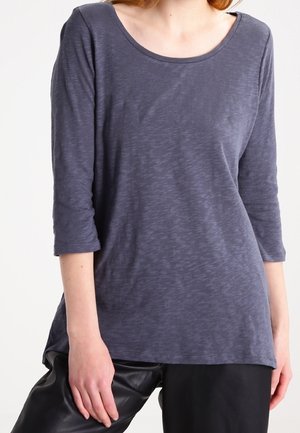 Femme portant un haut texturé gris foncé à manches longues et un pantalon noir, debout devant un fond clair uni.