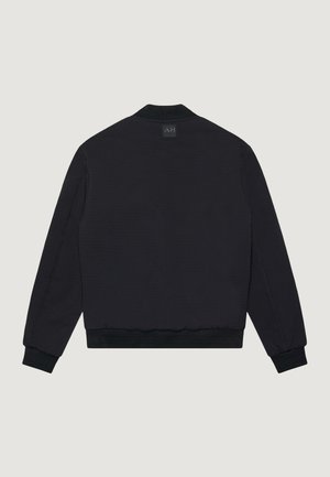 Giubbotto bomber nero a maniche lunghe con polsini e orlo a coste, visto da dietro, con una piccola patch rettangolare con logo sotto il colletto.