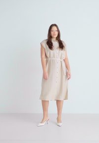 Vestido de linho bege com mangas curtas, fecho com botões à frente, cintura cingida por um cinto, comprimento até ao joelho, combinado com sapatos brancos de salto. Textura e design simples.