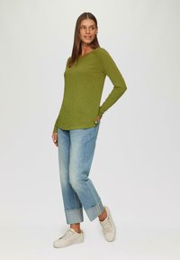 Olivgrünes langärmeliges Oberteil mit Rundhalsausschnitt, kombiniert mit hellblauen hochgekrempelten Jeans und beigen Sneakers, vor einem neutralen Hintergrund stehend.