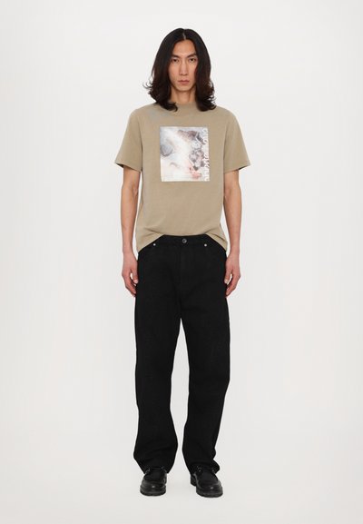 Camiseta beige de manga corta con un diseño cuadrado impreso, combinada con pantalones negros de corte holgado y con un patrón texturizado.