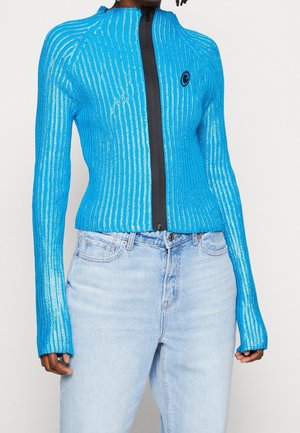 Blue ribbed knit jacket with long sleeves and a high collar, featuring a black front zipper and a small circular logo on the chest.